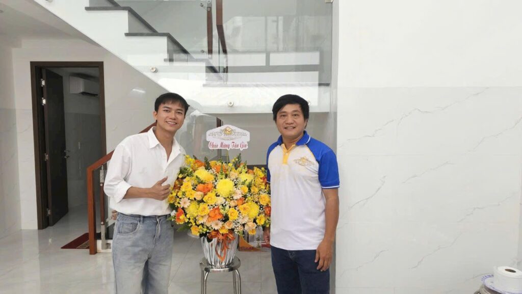 Hai người đứng cạnh lẵng hoa chúc mừng tân gia trong ngôi nhà hoàn thiện, phía sau là cầu thang kính.